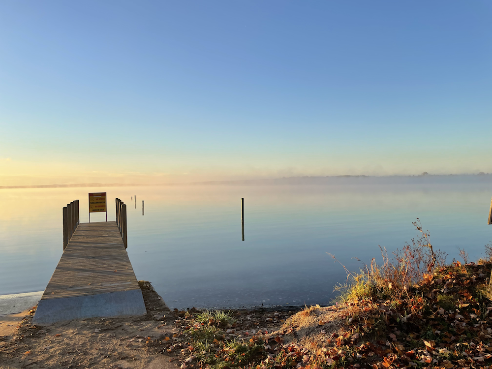 A dock jutting out into a lake. The sky is pink and yellow and blue, and those colors reflect onto the water. A light fog sits on top of the water.